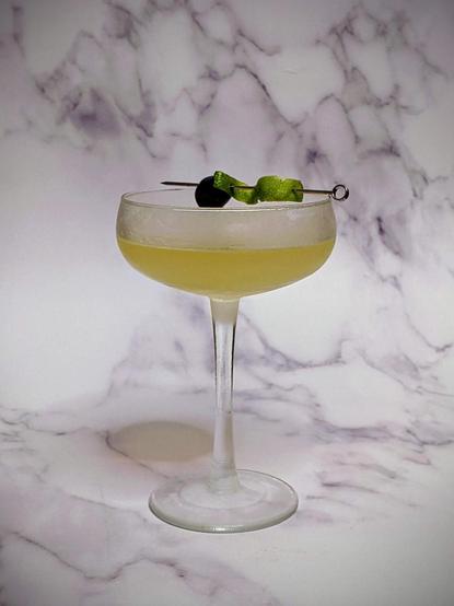 Chilled coupe glass with pale green drink and a cocktail pick with lime peel & cherry on the rim