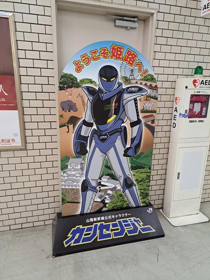 Cartel de bienvenida en la estación JR de Himeji.

Un personaje con armadura, en tonos azules y grises, y un casco. Arriba, en japonés, en caracteres de color naranja, está escrito el texto 