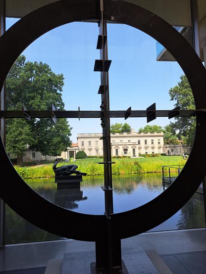 At the Virginia Museum of Fine Art in Richmond, looking through an open circle sculpture out the window to a small human-made water feature in which another sculpture, a human body in twisted form, rests. In the background are trees, blue sky, and another museum building. 