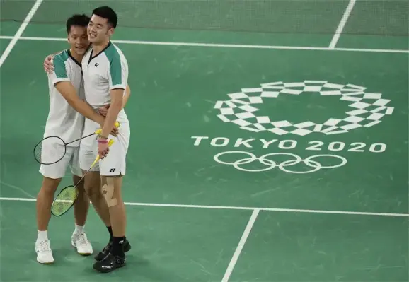 Taiwanese badminton players Lee Yang 李洋 and Wang Chi-lin 王齊麟 at the Tokyo 2020 Summer Olympics. The two are standing next to each other. Lee is hugging Wang with both arms around Wang's waist, while Wang has his arm over Lee's shoulder.