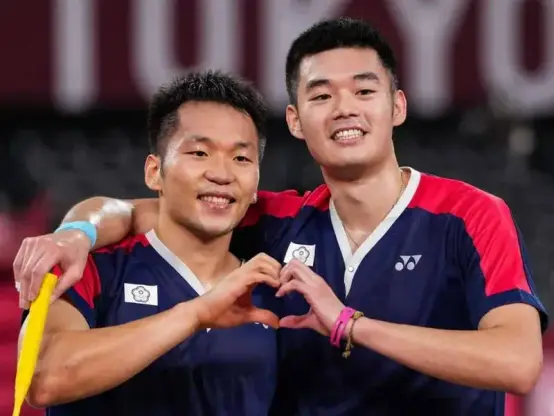 Taiwanese badminton players Lee Yang 李洋 and Wang Chi-lin 王齊麟 at the Tokyo 2020 Summer Olympic games. They are standing shoulder to shoulder, each of them holding up a hand, which are joined together to form a heart shape.
