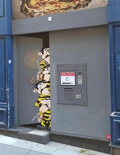 Streetartwall. A large, funny comic mural in the form of a paste-up has been installed on a double door of a former bank in the city center. It features four almost identical-looking but differently sized men with moustaches and yellow and black striped prison suits. They are the Dalton brothers from the Lucky Luke comic series. The door is slightly open and they are peering furtively around the corner (the open door is also drawn). Next to it is an ATM in the wall with a shop sign on the display and the words 