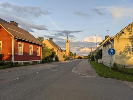 Un quartier pavillonnaire de Tampere, Finlande. Au centre une route, de part et d'autre des maisons en bois colorées. Au fond, une église. Il y a des arbres un peu partout. Le ciel est bleu, quelques nuages, lumière de soleil couchant.