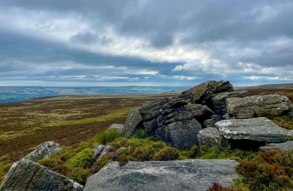 A crag of enormous, lichen-covered, grey sandstone boulders in a sea of bilberry shrubs. Behind, moorland sloping gently towards a valley of green fields and trees, all under a dramatic sky of blue-grey clouds.