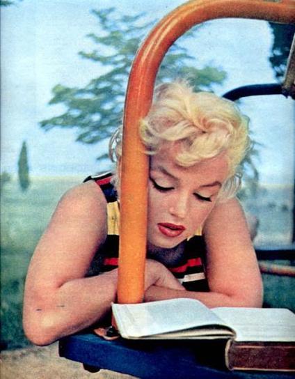 Photo of Marilyn Monroe, a blonde woman wearing a striped blouse, seen from chest up. Outdoor setting with trees in background. Monroe is leaning over supported by her elbows on a park table or bench, looking down at a book she is reading. 
