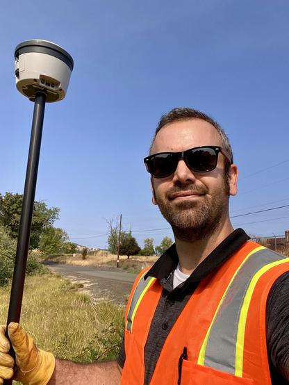 Me wearing a high-visibility orange vest and sunglasses, holding a surveying instrument in an outdoor setting with a clear blue sky and vegetation in the background.