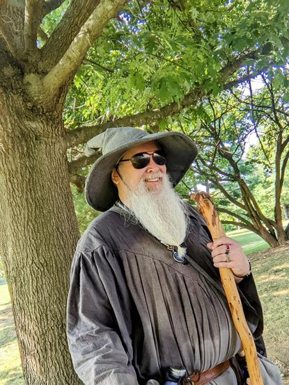 middle aged man with long grey beard in Gandalf style outfit with hat, staff, & sunglasses smiles at the camera underneath trees in the park
