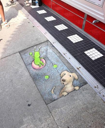 Streetart. On a gray sidewalk, a green monster (named Sluggo) and a brown dog (Farley) have been drawn on the ground with chalk. The two of them are sitting in a pool of water that has been painted on a long floor tile. Sluggo is lying in a pink swimming ring and has thrown two tennis balls with his short arms for the dog to catch. The dog looks out of the pool and looks around questioningly. Title: 