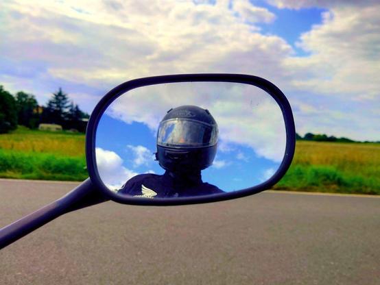 a rear view mirror of a motorcycle, a person with helmet is visible in the mirror. behind the person is blue sky with clouds. In front of the mirror is a road and a crop field. 