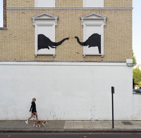 Streetartwall. Two black elephant heads have been sprayed into the white, closed window openings on the façade of a two-storey building in Chelsea. The two elephants are facing each other, almost touching with their trunks (the photo shows a passer-by walking her beagle in front of the building).
Note: A context has not yet been published. 05.08.2024, but there are definitely two elephants in the room.
