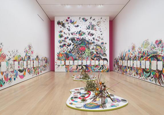 Gallery with walls drawn all over with colorful, semi-abstract animal and floral motifs. The walls are lined with white squares, some filled with individual drawings. On the floor are curving platforms holding abstract stick sculptures and bowls containing various materials
