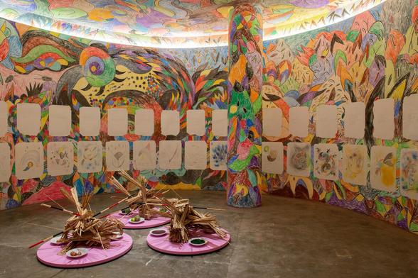 Gallery with a curving wall, pillar, and ceiling drawn all over with colorful, semi-abstract animal and floral motifs. The walls are lined with white squares, some filled with individual drawings. On the floor are three pink circles holding wooden driftwood sculptures