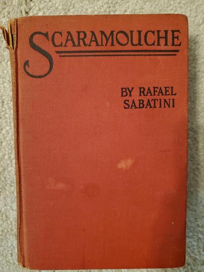 a red and waterspotted book cover, the 1921 edition of Scaramouche: A romance, by Rafael Sabatini. I read this darned thing to pieces...