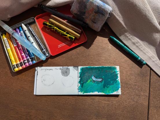 a top down photo of a small watercolor sketch of a sun chair with dappled shadows.  watercolor pastels and pens surround the drawing.