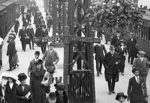 Photo looking down the platforms at Belfast's Queen's Quay railway terminus in early 20th century. Many well-dressed people make their way about their business. A sign in middle of the photograph indicates the left platform is for Bangor, the right hand platform is for Castlewellan