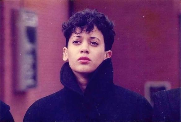 Amazing photograph of Kamala Harris in probably the 80s, looking so intensely, impossibly cool with short, dark, cropped hair, shaggy bangs, and black jacket with a large upturned collar. 