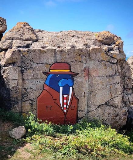 Streetart. The mysterious mural of a well-dressed walrus was painted on a stone in the dunes of a Breton beach. The blue walrus can be seen up to its chest, has two long white teeth and is wearing a reddish-brown suit with a matching hat with yellow stripes. He also wears a red and white tie. From this hidden vantage point, it has a good view of all the dark figures.
Title: 