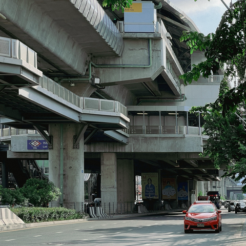 A modern urban scene showing a concrete elevated train station structure above a road with light traffic. A red car is prominent in the foreground, and lush green foliage can be seen alongside the road. The station appears to be part of a mass transit system, with signs indicating its name and usage.