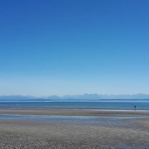 in the foreground, a beach at low tide with small areas of puddles. You can barely see a solitary walker and a person on a paddle board.  In the distance, island and beyond them, the Coast mountains disappearing in the haze of a hot summer day. The sky is blue, not a cloud in sight. 