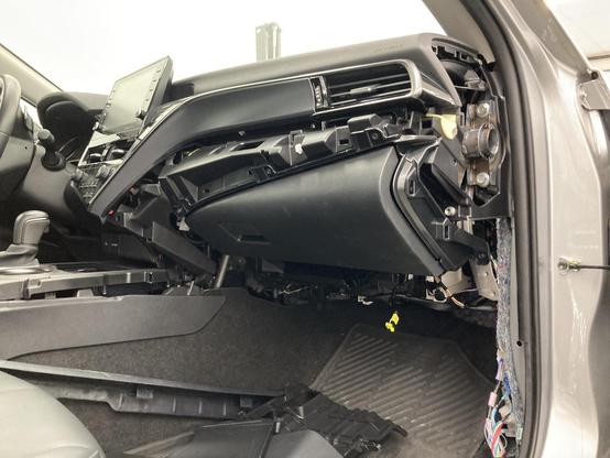 A disassembled glove compartment of a 2023 Toyota Camry. Much of the trim panels have been removed around the actual glove box. Below the glove box is a yellow plug, where the airbag used to be connected to. Exposed wires can be seen along the passenger side A column.