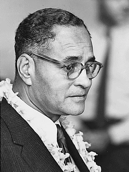 Photo of Ralph Bunche looking to the right.  He is wearing eyeglasses, dress jacket, white shirt, and tie with a garland of flowers around his neck.  