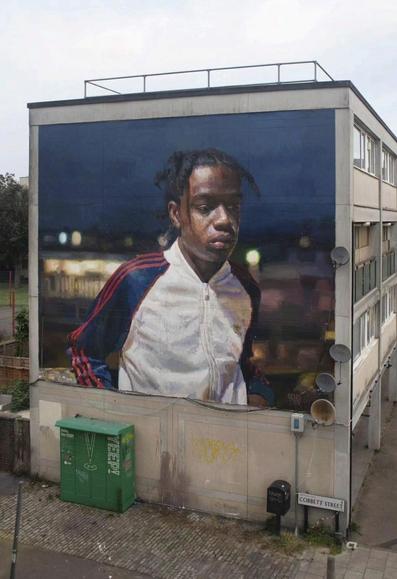 Streetartwall. A magnificent mural of a young black man with a short Rasta hairstyle against the night-time backdrop of Brixton was sprayed/painted on the outside wall of a two-storey block of flats. The mural is photorealistic and shows the British rapper Loyle Carner in a white jogging jacket with red and blue sleeves. In the background is a dark evening sky and the blurred lights of a train station. As always, a fantastic work by the artist in a rather bleak area.
Info: The title is taken from the song of the same name by British rapper Loyle Carner.