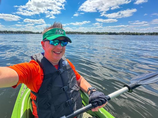 Person kayaking on a lake, wearing an orange shirt, life vest, green visor, and sunglasses, with blue sky and scattered clouds in the background.