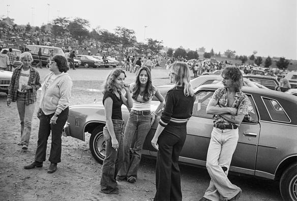 Black and white photo, 70s: Young people standing around in a lot full of cars next to a park.

Photo by Don Hudson