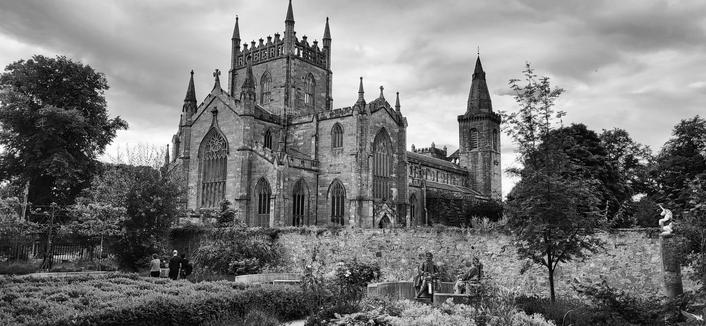 Black and white photo of the Dunfermline New Abbey in Fife,  Scotland,  photo shows in the foreground a small walled garden with two sitting statues of gentlemen awaiting some company. The sky is cloudy and moody looking.