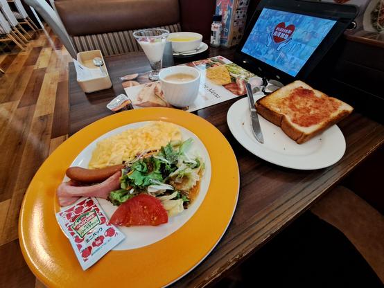 Foto de un desayuno de estilo occidental en un restaurante cercano al hotel, con ensalada, una salchicha, bacon, una tortilla, sopa de maíz, yogur con frutas, una tostada con mantequilla y mermelada de fresa y un café con leche. Se ve también la tablet desde la que se realizan los pedidos.