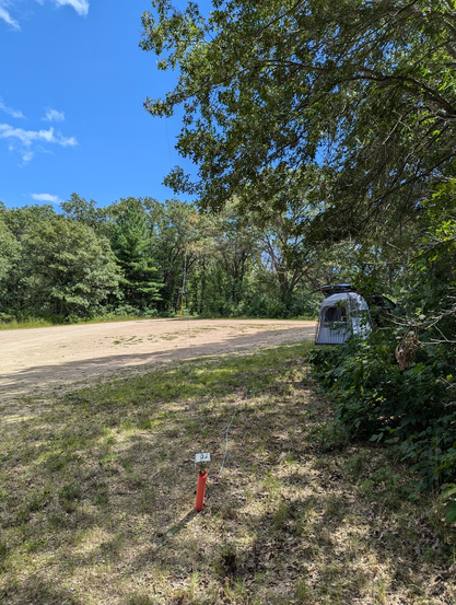 A view of a dirt parking area.
The far side of the dirt lot is lined with trees, along with the right side.
The sky is mostly blue, with a few wispy clouds.
The ham pod is near the right side, in the middle distance.
The ground near the bottom has some scrubby grass, and the red post with the antenna.
The antenna goes up over a branch of a tree, and to the yellow pole and red post on the far side of the lot.