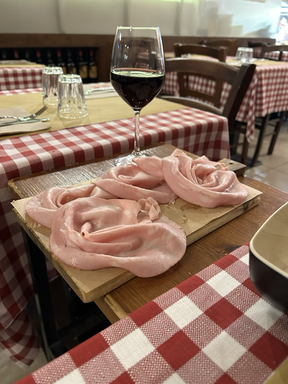 A wooden board with slices of bologna and a glass or fed wine. In the background a table with one place setting and a red and white checked cloth