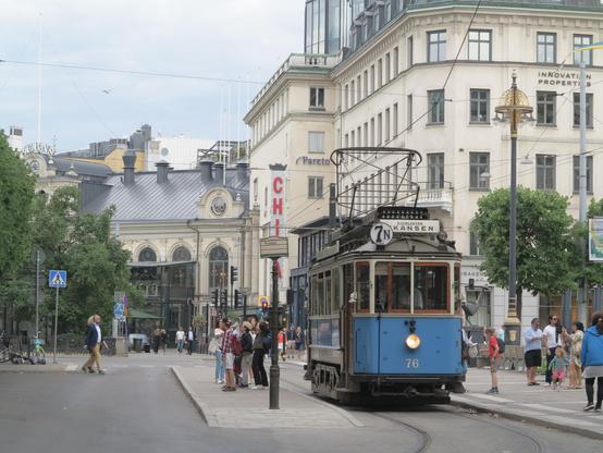 An historic 4-wheeled electric tram in a primarily light blue livery sits at a tram stop on a single track loop line with a low platform on each side.  In the background is a series of multi-storey grand 19th century commercial buildings.