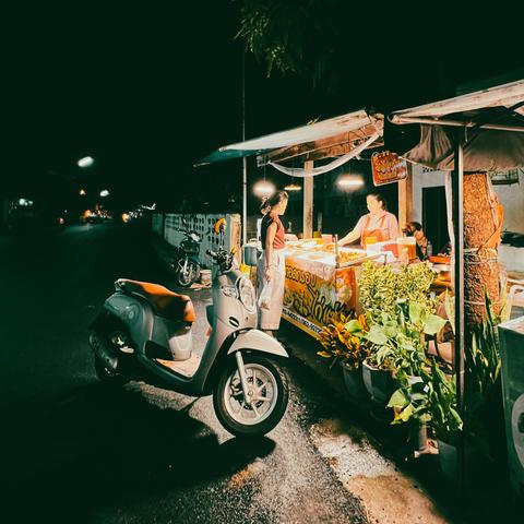 A night scene of a small street food stall in Hat Yai, Thailand, illuminated by warm lights, set against a dark street. A scooter is parked in front of the stall, where a customer is speaking with the vendor. The stall is surrounded by potted plants, adding a touch of greenery to the cozy ambiance, reflecting the local culture and night market vibe.