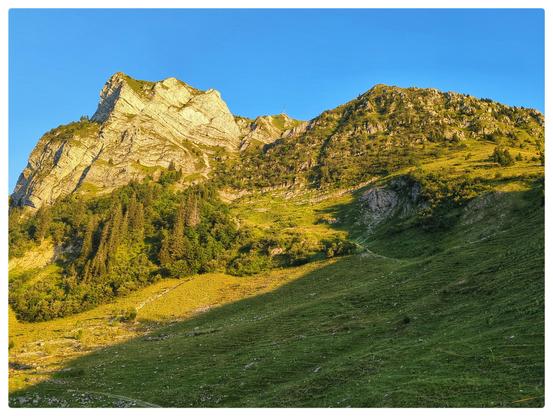 Ein Berge erhebt sich von einer Alpwiese, beleuchtet im Abendlicht.