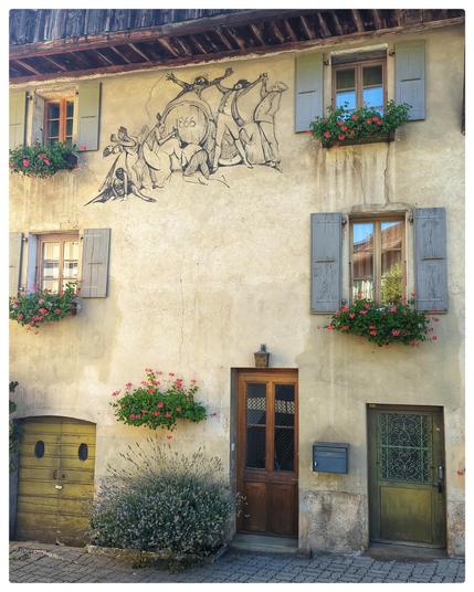 Hausfassade, Blumen an den Fenstern. Unter dem Dach ist die Szene einer Party von 1866 aufgemalt.