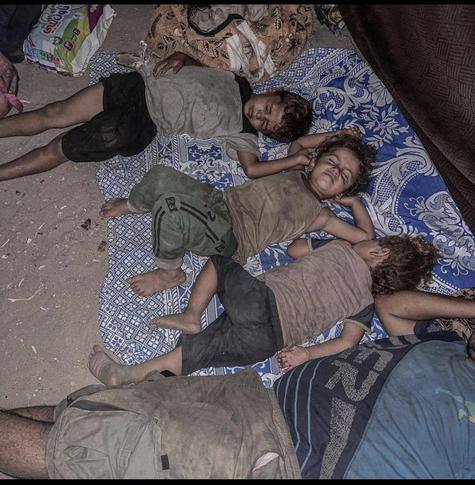 Three war torn children sleep on ground and covered in dirt wearing tattered clothing. 