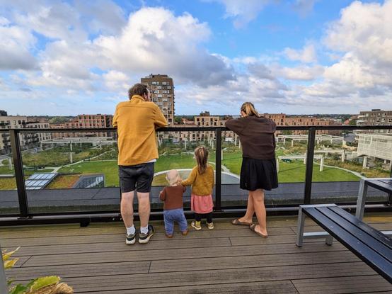 Photo of four people standing at a glass railing on the top of a roof, with their backs turned to the camera. They are looking at a green park and apartment buildings. The four people, from the left, an adult man, a very small/baby body, a toddler girl, and an adult woman.