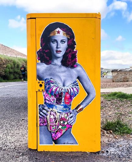 Streetart. A cool, self-designed paste-up of actress Lynda Carter as Wonder Woman was created in pop art style and stuck as a poster on a yellow distribution box on the beach. Her face and body are in purple with colored elements. Wonder Woman has red, long, wavy hair and wears a golden headband with stars on her forehead. She is wearing a skimpy bodysuit without arms with glitter elements and a belt. With her hand on her hip, she looks directly at the viewer. 
Info: The poster was pasted on the occasion of the 72nd birthday (24.07.1951) of the actress Lynda Carter.