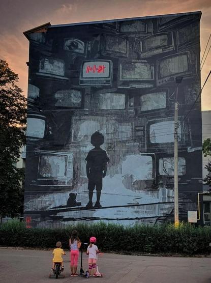 Streetartwall. The impressive mural of a young boy in front of a wall of stacked televisions was sprayed/painted on the exterior wall of a four-story building. The mural is mainly in white, black and shades of gray. A little boy stands with his back to us in front of a huge wall of televisions, and next to him lies a teddy bear. Soon he will no longer be playing, but staring at the stories on the sets. On one TV set, the following appears in red: 