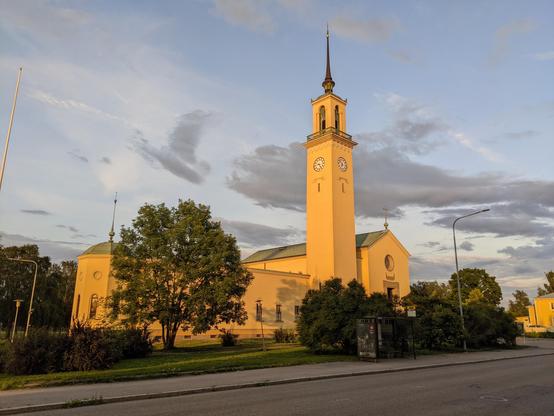 Une belle église de Tampere en Finlande. Les murs sont jaunes, on voit un clocher carré, haut et fin, au centre. Il y a des arbres devant l'église. Le ciel est bleu avec quelques nuages. Lumière de soleil couchant. Au premier plan, une route et un abribus.