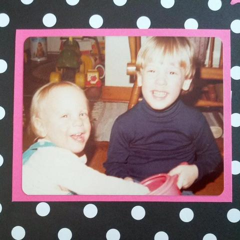 Two young children are smiling and playing indoors. The child on the right, wearing a dark turtleneck sweater, holds a red object. The child on the left, wearing a light shirt with blue overalls, leans towards the other child with a joyful expression. The background shows a carpet, a wooden chair, and a few toys. 