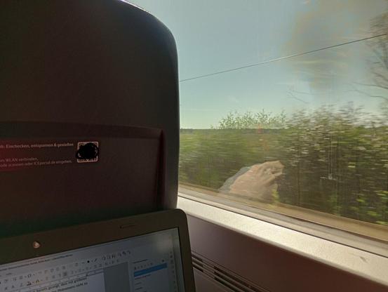 Foto vom Sitzplatz am Fenster aus. Draußen sieht man die Landschaft bei strahlend blauem Himmel. In der unteren linken Ecke des Bildes ist der Bildschirm des Laptops mit geöffneter Textverarbeitung zu sehen.