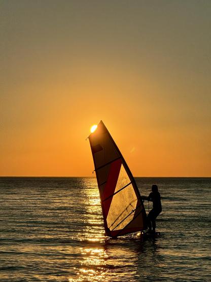 A windsurfer on the sea, holding the surf sail and the sun is about to set behind the top of the sail. Sky is a gradient shades of orange and the sea is dark blue.