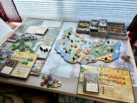 A game of Spirit Island is set up, featuring a game board with various colored pieces, cards, and tokens. Organizers are filled with additional game components. The game appears to be in progress with several elements strategically placed. All of the components and boards are on a tray in front of sunlit closed blinds.