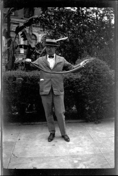 Black & white photo of a man dressed in early 1920s style of a suit, bow-tie, and a straw boater hat standing in a park.  He holds a snake over a meter long in his hands, his arms extended to either side.  Behind him is seen park shrubbery and further back a bit of a multi-story stone building. 