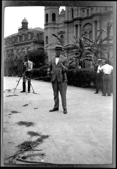 Black & white photo of a man dressed in early 1920s style of a suit, bow-tie, and a straw boater hat standing in a park.  He holds a snake over a meter long in his hands, one end of the snake dangling down; it might be dead. To left stands another man with a camera mounted on a tripod. In background are a church and a multi-story stone building, New Orleans' Saint Louis Cathedral and Cabildo. 