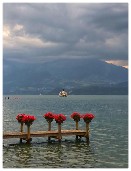 Ein Steg am Ufer eines Sees. Auf den Stützpfosten sind Blumen arrangiert. Ein Kursschiff nähert sich. Am anderen Seeufer Berge, die von dunklen Wolken verdeckt sind.