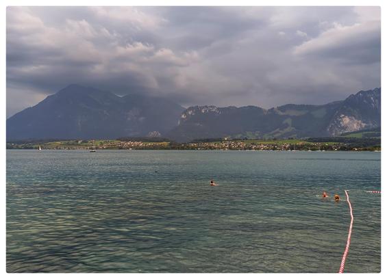 See mit klarem Wasser, einige Schwimmer. Am anderen Seeufer Berge mit dunklen Wolken. Der See ist im fahlen Abendlicht eines nahenden Gewitter.