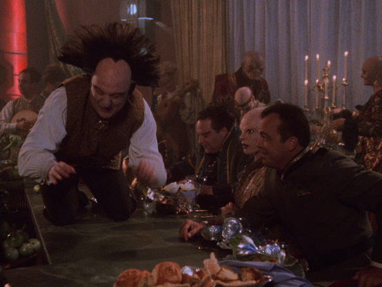 A frame from the first episode of Babylon 5 showing a drunk Londo sitting on his knees on a long table full of food, waving his arms, while a number of people are sitting along the table watching him.

The image has a lot of fine film grain and digital noise from compressing the film grain.